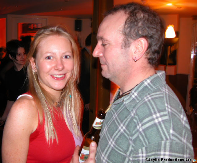 A smiling woman in a red top stands beside a man in a check shirt holding a beer bottle