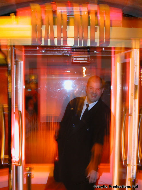 A man in a dark suit stands in a doorway under a lit Bar Med sign