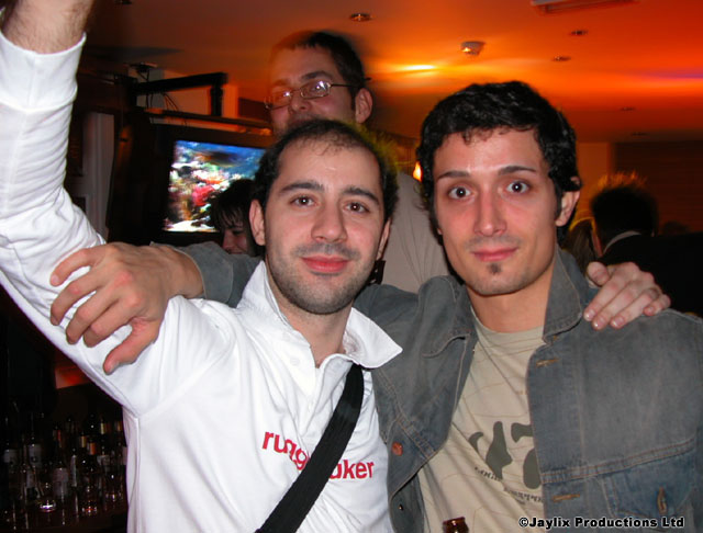 Two men pose with arms around each other, one in a white top and one in a denim jacket