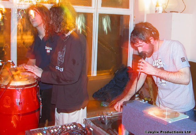 A man with headphones sings into a microphone at a DJ desk beside a red conga drum