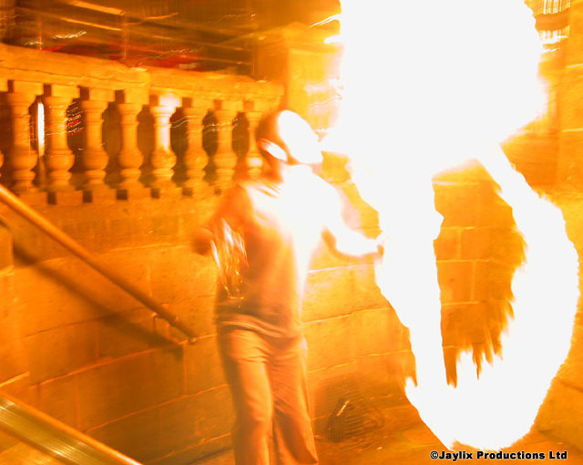 A blurred person stands beside a large flame near a balustrade breathing fire