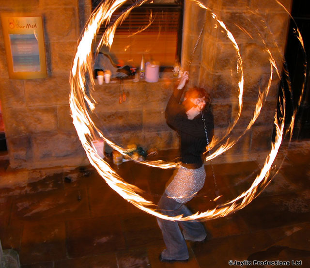 A person spins a circle of fire poi in front of a stone wall at night