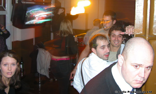 A group of people crowd together, a bald man in black in front, with a television behind