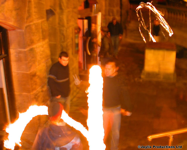 People stand on a stone terrace at night with tall flames and fire trails