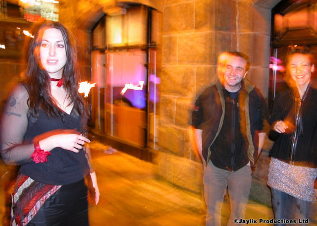 Three people stand outside a stone building at night, a woman in black on the left
