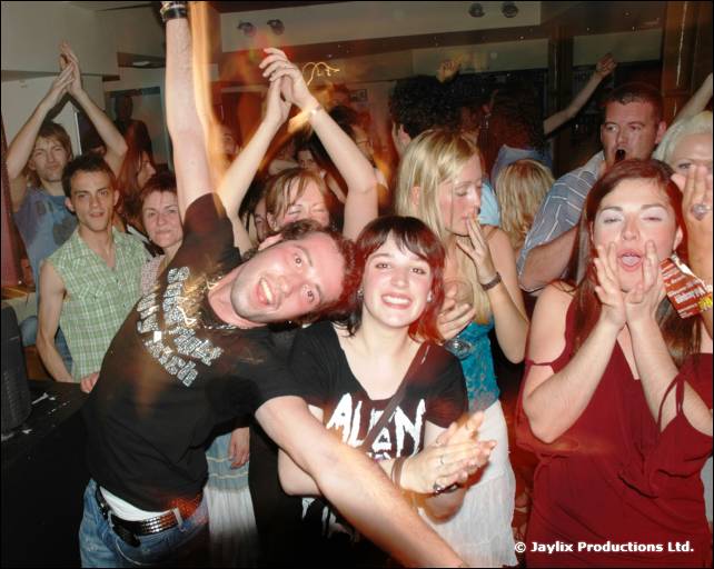 Crowd with raised arms, including a man leaning forward in a black T-shirt, a woman at his shoulder, and a woman in red shouting