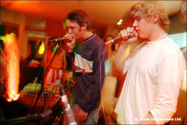 Two beatboxers with microphones, one in a navy top and one in white, with a flame effect behind