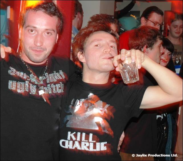 Two men pose, one in a black shirt and one in a Kill Charlie T-shirt drinking from a glass