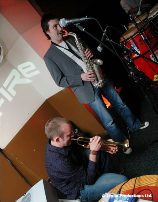 Saxophonist standing and trumpeter seated play beside a FIRE sign, with drums in the corner