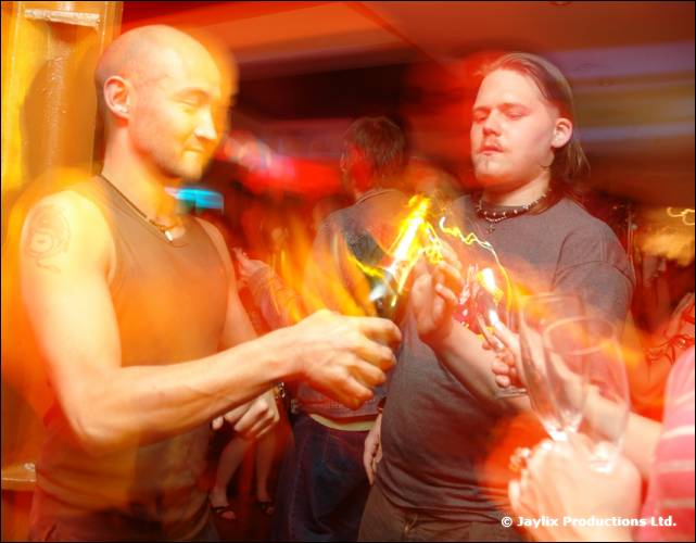 Two men in vests open a champagne bottle among blurred movement, with glasses held out