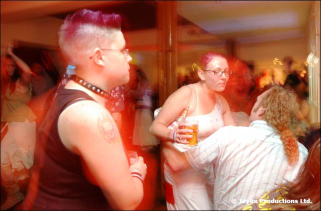 Person with pink spiked hair and a studded collar stands in a bar, with two people dancing beyond