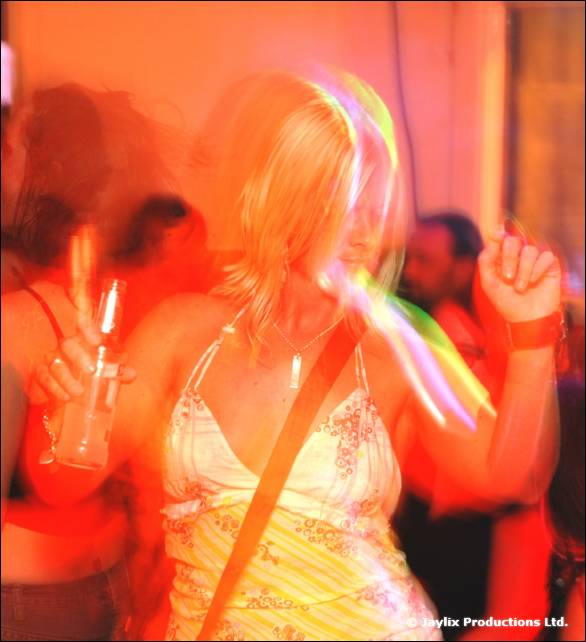 Blurred woman in a yellow patterned top holds a bottle and dances, with glow stick trails