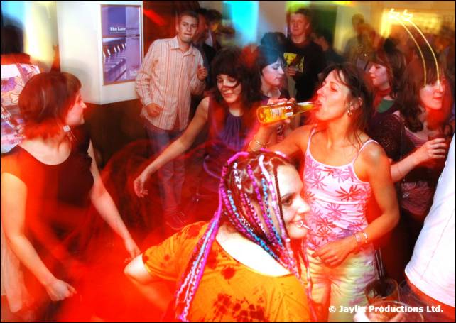People dancing in a busy bar, a woman in a pink top tilting a bottle to her mouth, another with coloured braids in the foreground