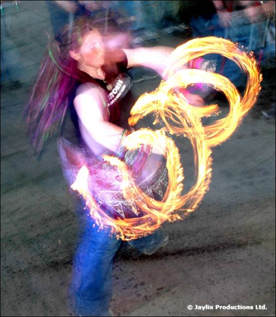 Person with long hair spins fire poi in looping trails on a paved area