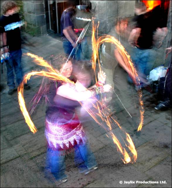 Person in a pink patterned skirt spins fire poi on a paved area, with people watching behind