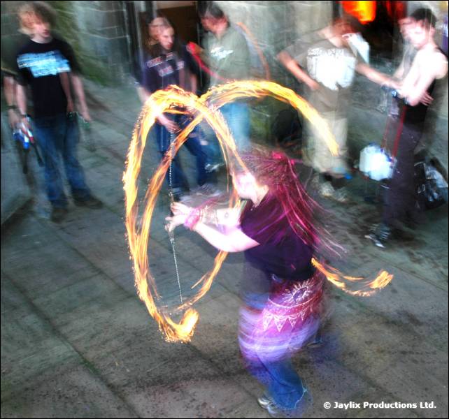 Person in a purple skirt spins fire poi on a paved area, with onlookers standing behind