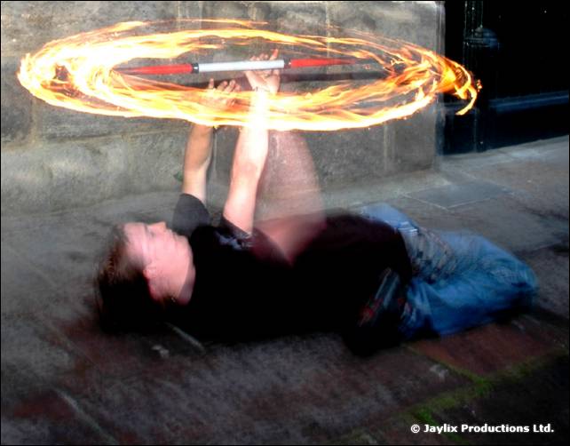 Person lies on their back on a paved floor, spinning a fire staff above them