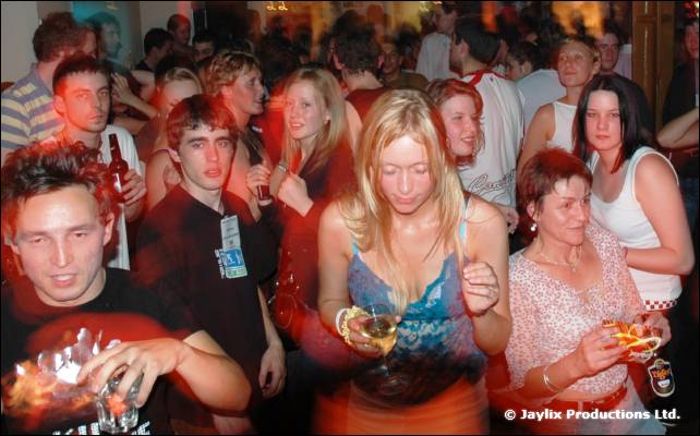 Crowd at a club night, with a blonde woman in a blue top looking down holding a drink, and others around her