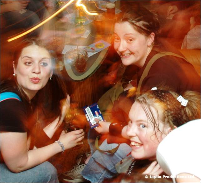 Three women crowd together in a blurred, orange lit photo, one holding a small card