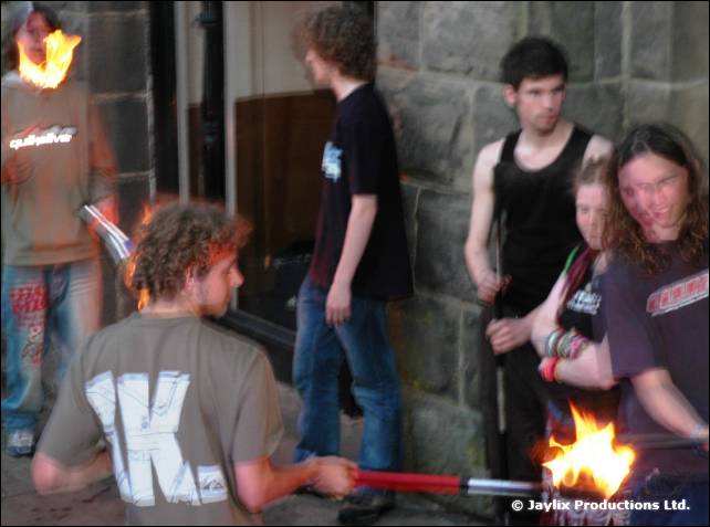 Person in a grey T-shirt lights a flaming staff, with several people standing by a stone wall