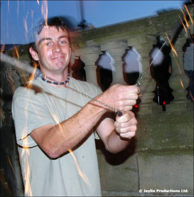 Smiling man in a grey T-shirt holds a chain and fire poi, in front of a stone balustrade