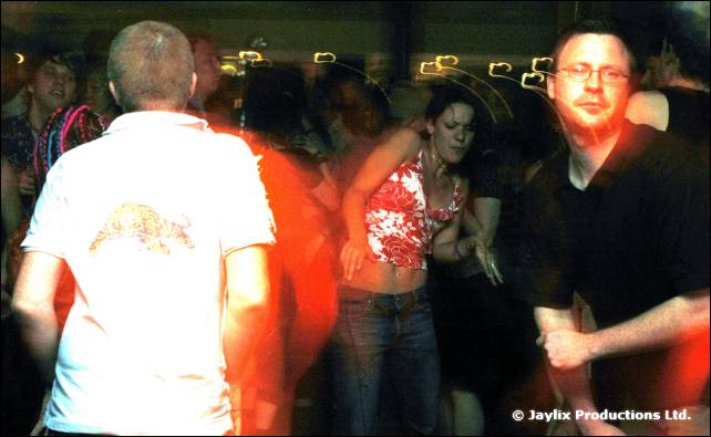 Dancers in a dark bar, with a man in glasses and a black shirt on the right and a man in white on the left