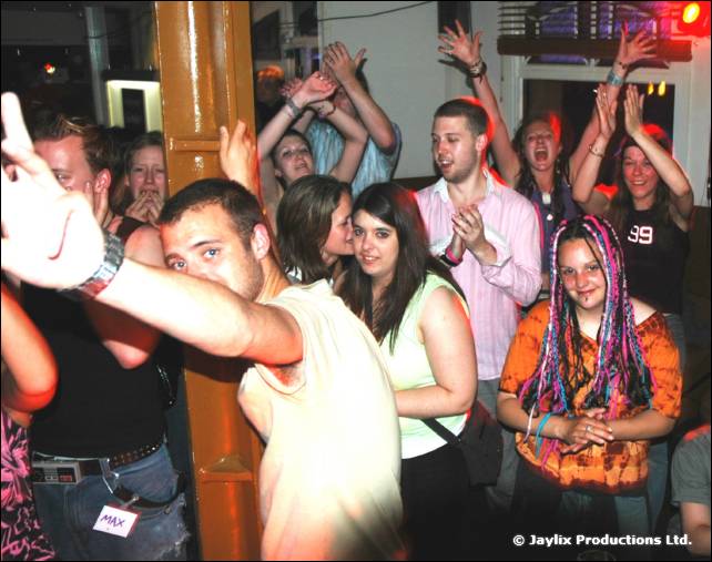 Crowd with raised arms dancing by an orange pillar, with a woman with pink and blue dreadlocks at the front