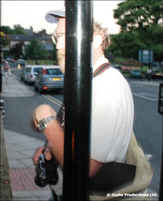 A bearded man in glasses and a cap stands behind a black post on a street, holding a camera