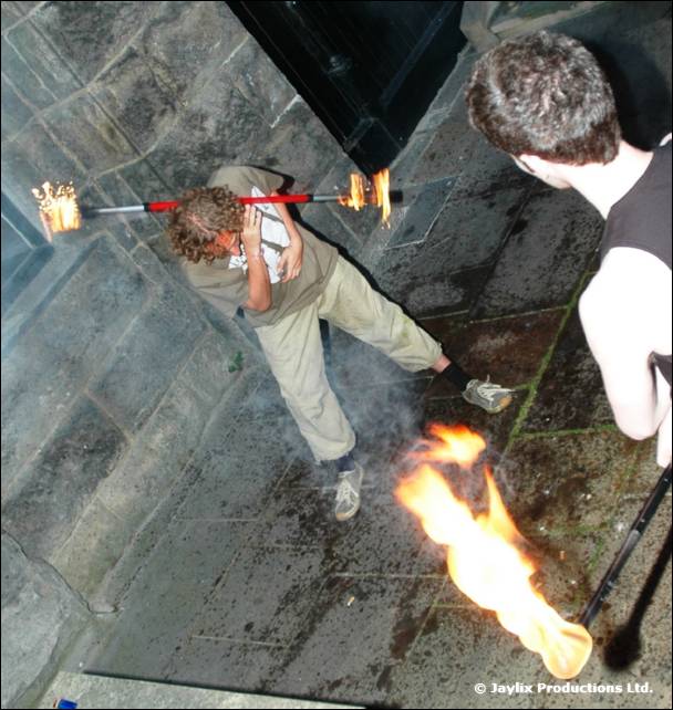 Person lights a staff with flaming ends over a wet paved area, watched by a person at right