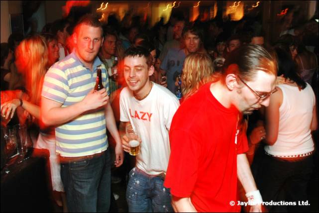 Three men in the foreground of a busy bar, one in a striped top, one in a white Lazy T-shirt and one in red