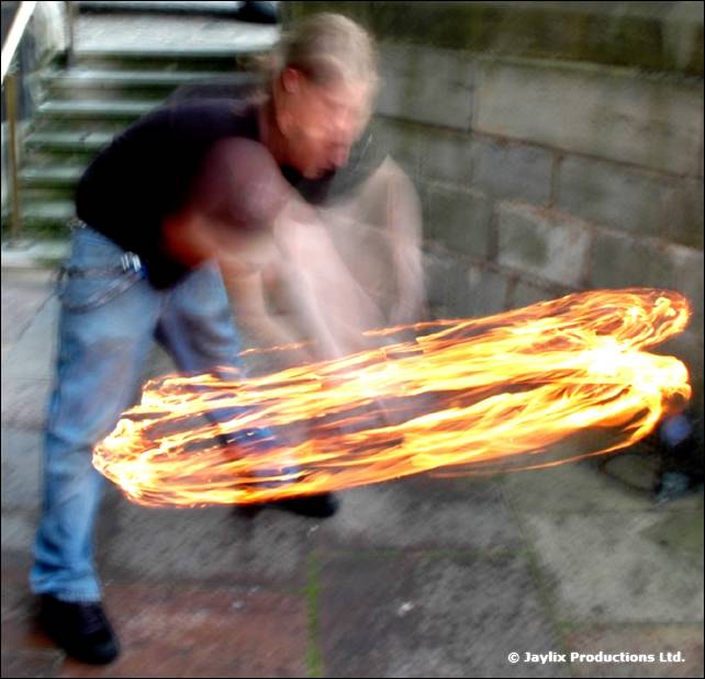 Person in a dark top and jeans swings a fire staff in a blurred circle, beside steps