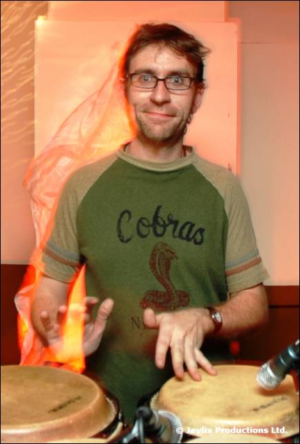 A smiling person in glasses and a green Cobras t-shirt plays congas, with a flame blurred behind