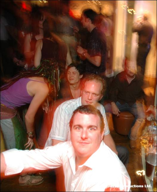 Two seated men in white shirts look at the camera among a blurred, dancing crowd