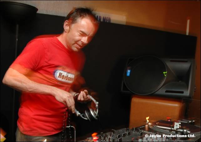 A DJ in a red t-shirt holds headphones beside turntables, with a speaker behind