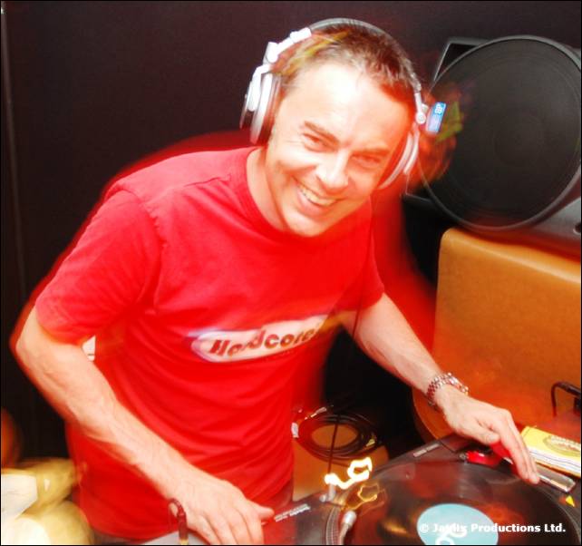 A smiling DJ in headphones and a red t-shirt leans over a turntable