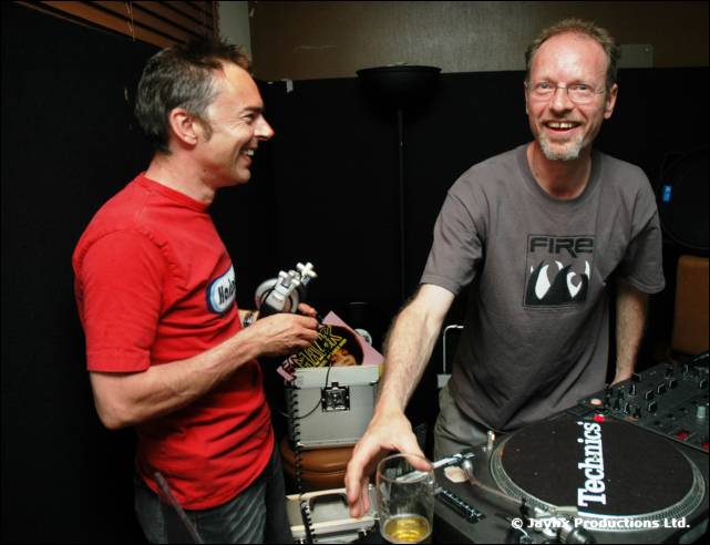 Two men laugh beside a Technics turntable, one in a red t-shirt, the other in a grey FIRE t-shirt