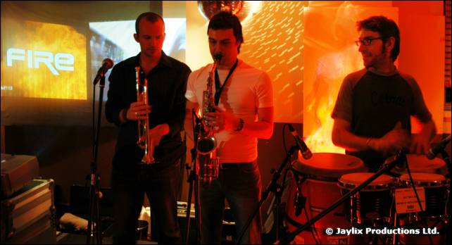Two people play saxophone and clarinet beside a drummer, in front of a FIRE projection