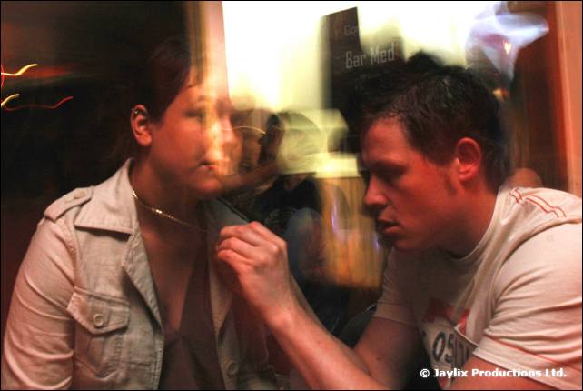 A person in a light jacket and a man in a white t-shirt lean close together, a bar sign behind