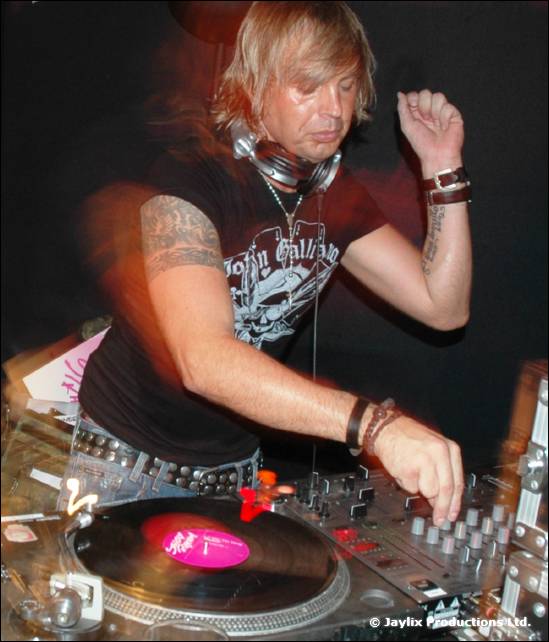 A DJ with blond hair and headphones around his neck mixes at a turntable and mixer