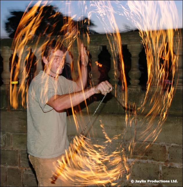 A person in a grey t-shirt swings a flaming chain outdoors at dusk, leaving fiery trails