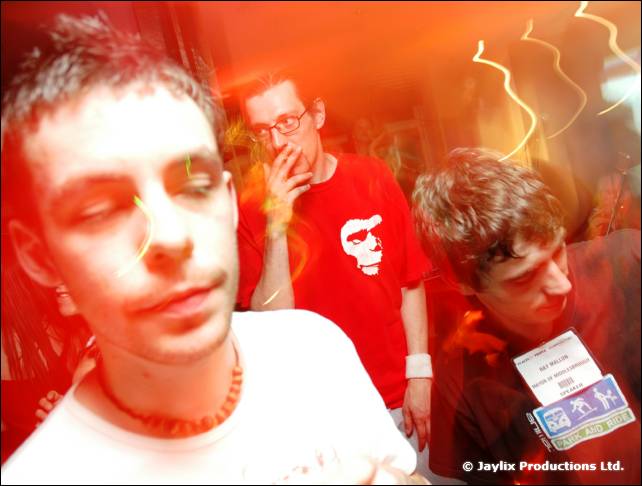 Three blurred people in a red-lit room, one with a wooden bead necklace and another in a red t-shirt