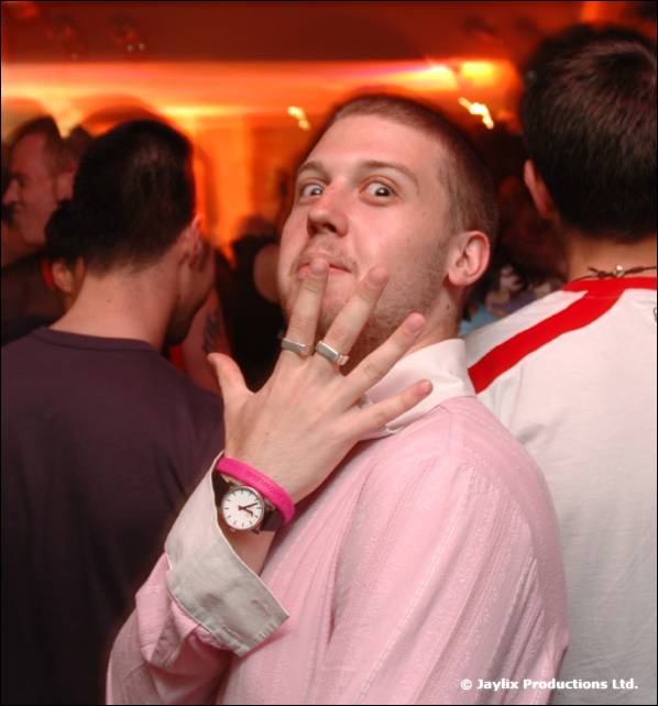 A man in a pink shirt holds two fingers to his chin and raises his eyebrows, a crowd behind