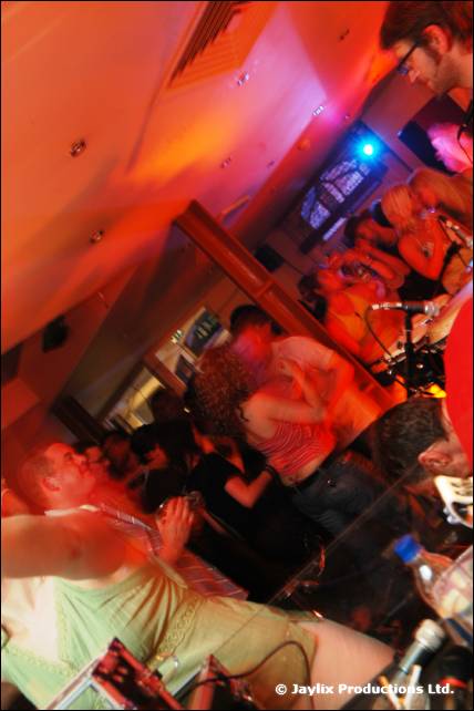 A tilted, red-lit view of people in a busy bar with a person in the foreground near equipment