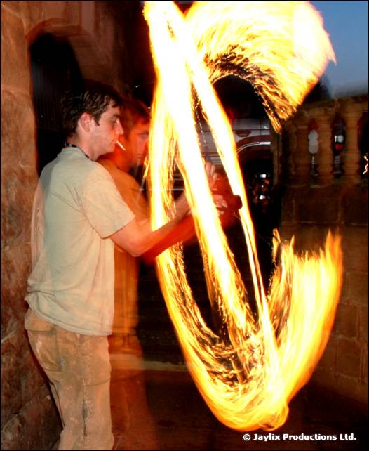 A person in a pale t-shirt spins fire poi beside a stone arch at dusk