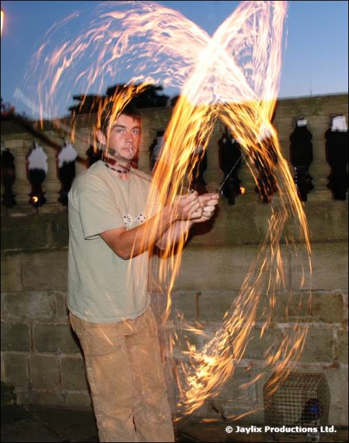 A person in a pale t-shirt spins fire in front of a stone balustrade at dusk