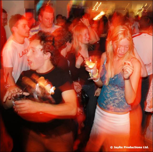 A dancing crowd in a bar, with a man in a black t-shirt and a blonde woman with a drink in front
