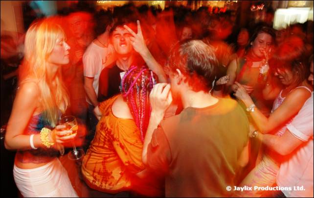 A blurred crowd dances in a busy bar, with a blonde person and someone with red braided hair in front