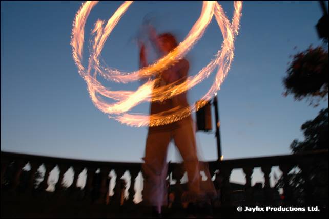 A blurred person twirls fire poi against a blue dusk sky in front of a stone balustrade