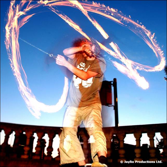A person in a grey t-shirt swings long trails of fire poi against a blue sky, with a balustrade below