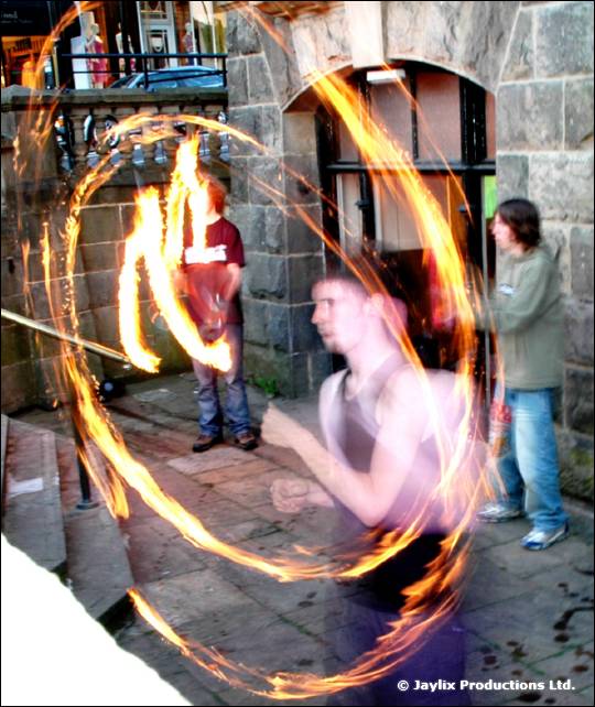 A person swings a fire staff in a street beside a stone archway, with two people watching